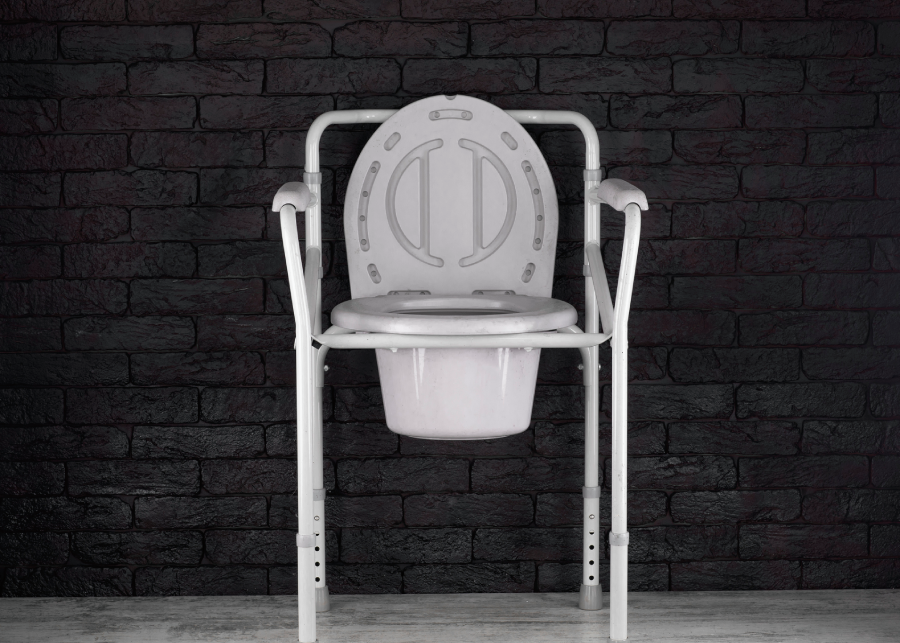 A white adult potty chair with armrests against a dark brick wall.